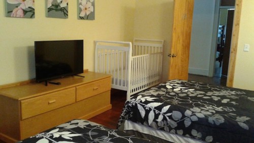 A cozy room with two beds, a TV on a dresser, and a crib in the corner. Floral decor on the walls.