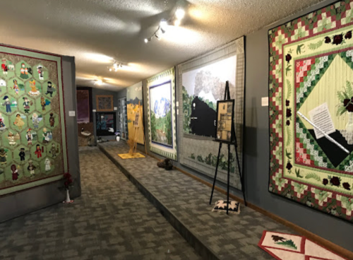 A gallery interior featuring colorful quilts displayed on walls, with a carpeted walkway and artwork on easels.