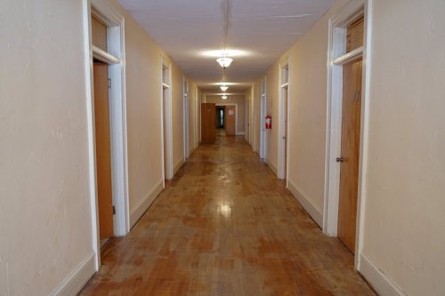 A long, empty hallway with wooden floors and doors on either side, illuminated by ceiling lights.