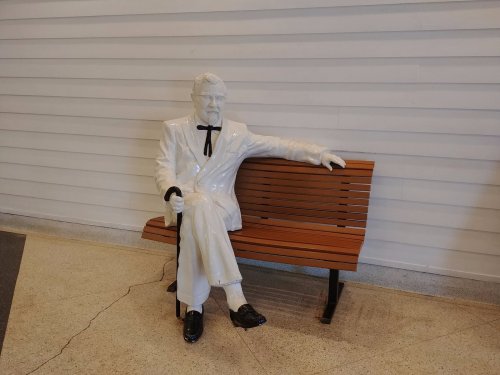 A life-sized statue of a man in a white suit sitting on a wooden bench, holding a cane.
