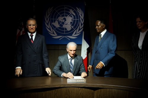 A man in a suit signs a document at a table, flanked by two figures, with a United Nations backdrop.