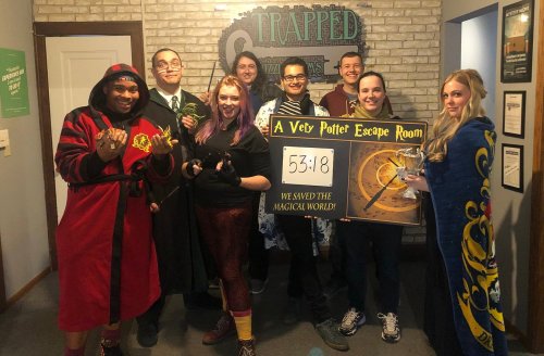 Group of eight people in themed costumes celebrating after completing a Harry Potter escape room, holding a sign with their time.
