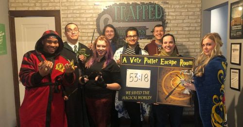 A group of eight people in themed costumes celebrate after completing a Harry Potter escape room, holding a sign with their time.