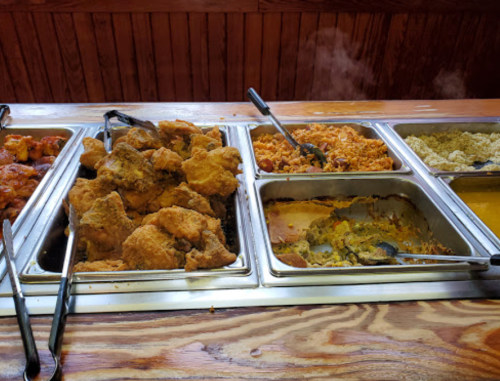 A buffet with trays of fried chicken, rice, and various side dishes on a wooden counter. Steam rises from the food.