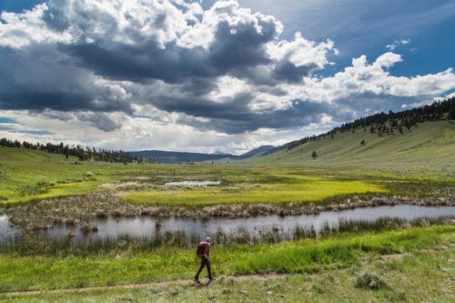 A hiker walks along a grassy path beside a serene pond, surrounded by rolling hills and dramatic clouds.