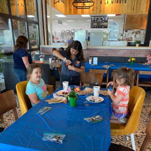 A restaurant scene with two children at a blue table, a server nearby, and other diners in the background.