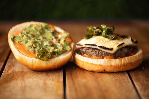 A burger with melted cheese and jalapeños on a toasted bun, next to a bun spread with guacamole.