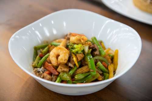 A white bowl filled with colorful stir-fried vegetables and shrimp on a wooden table.