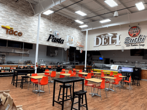 A spacious food court with tables and chairs, featuring signs for taco, pasta, deli, and sushi stations.