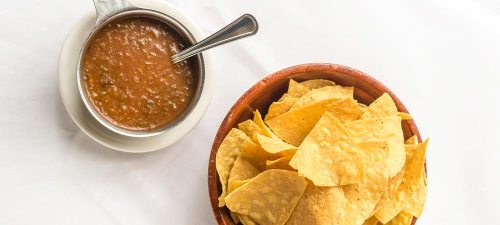 A bowl of crispy tortilla chips next to a small dish of salsa on a white tablecloth.