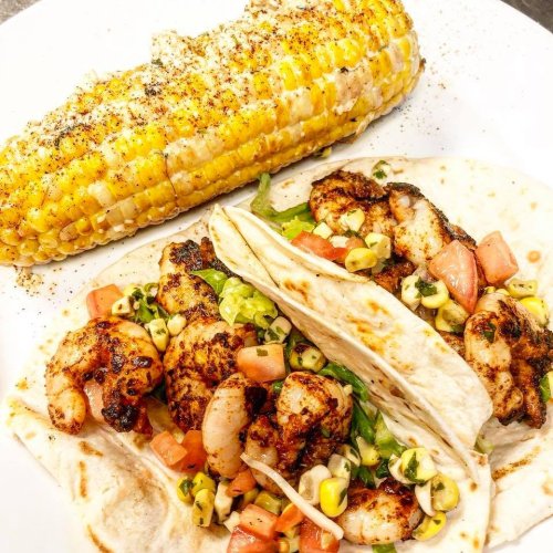 Two shrimp tacos filled with lettuce, tomatoes, and corn, served with a side of grilled corn on the cob.