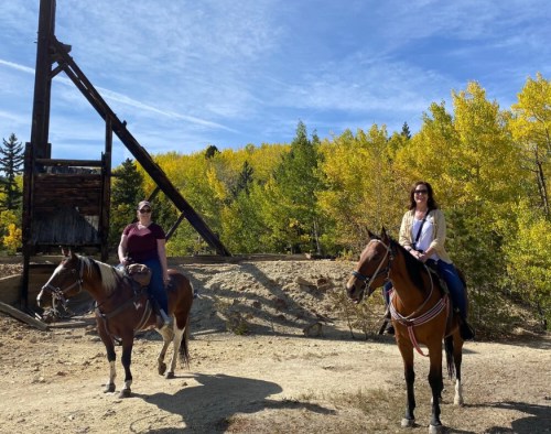 Two women on horseback in a scenic outdoor setting with autumn foliage and a wooden structure in the background.
