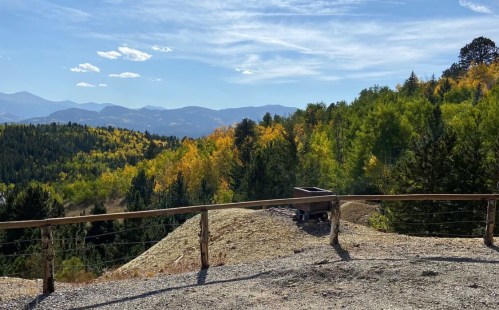 Scenic view of rolling hills with autumn foliage, mountains in the background, and a wooden fence in the foreground.