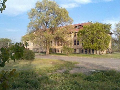 An old, two-story building surrounded by trees and greenery under a clear blue sky.