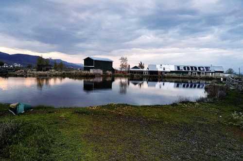 A tranquil lodge by a calm pond, surrounded by greenery and mountains under a cloudy sky.