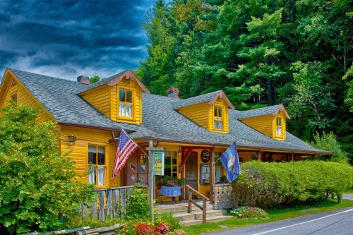 A charming yellow house with a porch, surrounded by greenery and flags, under a dramatic sky.