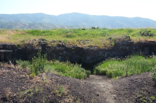A rocky entrance to a cave surrounded by tall grass and mountains in the background under a clear sky.