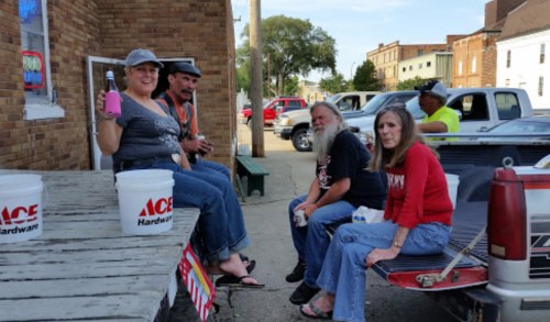 A group of people sitting on a bench and truck tailgate outside a hardware store, chatting and enjoying drinks.
