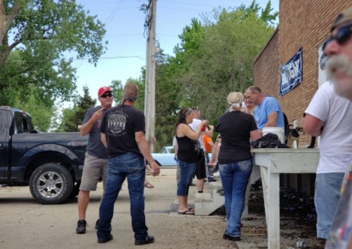 A group of people socializing outside a building, with a truck parked nearby and trees in the background.