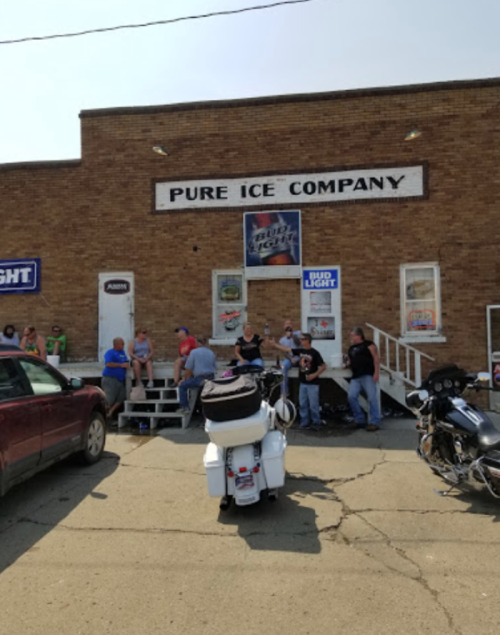 A brick building with "Pure Ice Company" sign, people sitting on steps, and parked motorcycles in front.