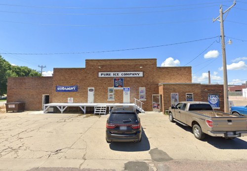 A brick building with "Pure Ice Company" sign, featuring a porch and parked vehicles in front under a clear blue sky.