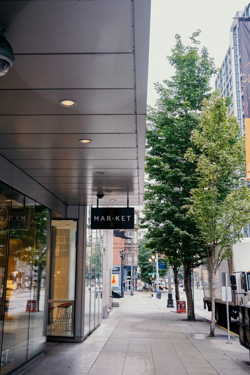 A city sidewalk with a sign reading "MARKET" and trees lining the street, creating a calm urban atmosphere.