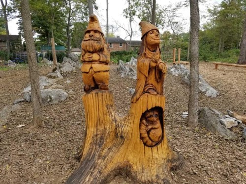Wooden carvings of a gnome and a woman on a tree stump, surrounded by rocks and trees in a park setting.