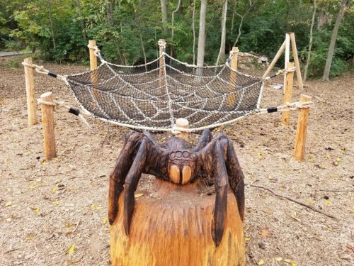 A wooden spider sculpture sits on a stump in front of a rope web structure in a forested area.