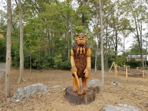 A wooden sculpture of a troll stands in a forested area, surrounded by trees and fallen leaves.