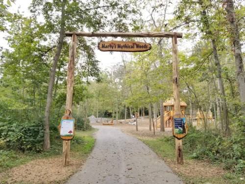 Wooden archway sign reading "Marty's Mythical Woods" at the entrance of a forested path with trees and play structures.