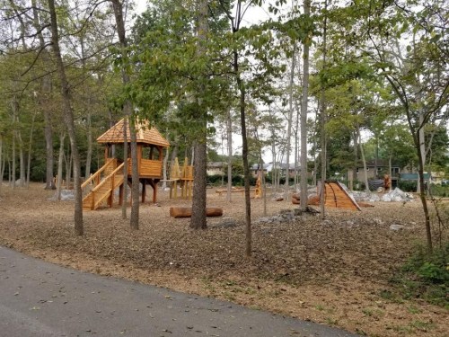 A wooden playground surrounded by trees, featuring slides and climbing structures, set in a natural, leaf-covered area.