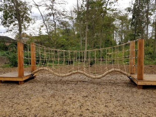 A rope bridge made of wooden posts and netting, set in a wooded area with a ground covered in wood chips.