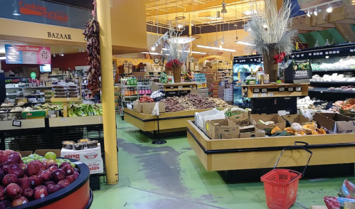 A vibrant grocery store interior with fresh produce, shelves of goods, and a shopping basket in the foreground.