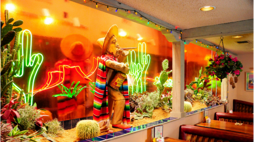 Colorful restaurant decor featuring a sombrero-wearing statue, neon cacti, and vibrant plants in a cozy setting.