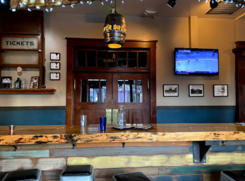 A rustic bar with a wooden counter, stools, and a TV showing a sports game, framed photos on the wall, and a ticket sign.