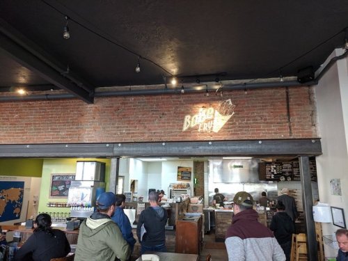 Interior of a casual eatery with a brick wall, customers waiting in line, and a menu visible in the background.