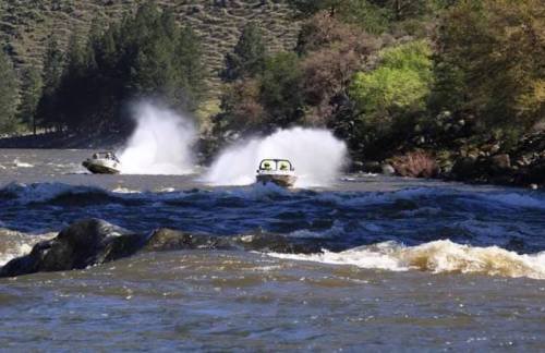 Two boats speed across a river, creating splashes against a backdrop of trees and rocky shores.