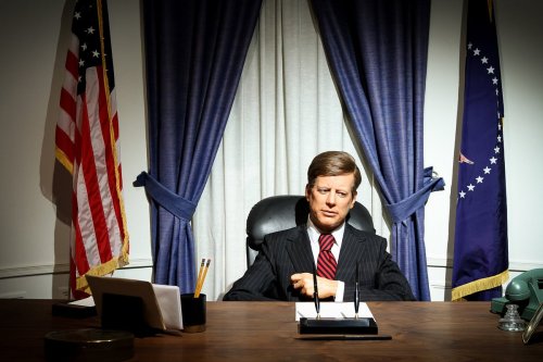 A lifelike figure of a seated man in a suit at a desk, flanked by American flags and curtains.