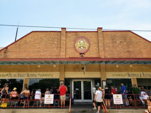 A bustling brewpub with a brick facade, featuring a sign for Pecan Street Brewing and a line of customers outside.