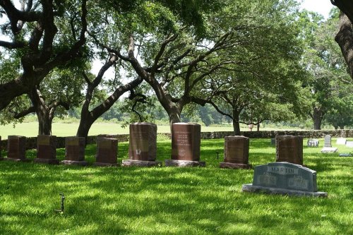 A peaceful cemetery with several gravestones under large trees, surrounded by lush green grass.