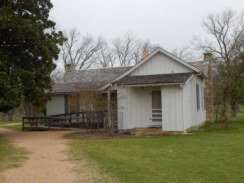 A small, white wooden house with a sloped roof, surrounded by grass and bare trees on a cloudy day.