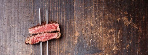 Sliced steak resting on a wooden surface, with two metal skewers positioned beside it.