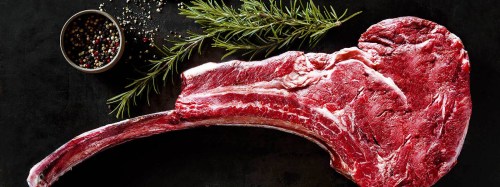 Raw tomahawk steak with fresh rosemary and a bowl of mixed peppercorns on a dark surface.