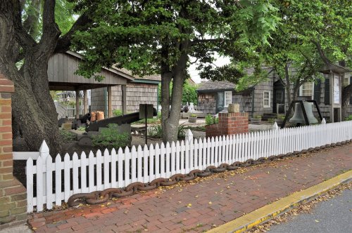 A quaint outdoor scene featuring a white picket fence, trees, and historic buildings in a charming neighborhood.