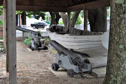 A wooden boat and two cannons are displayed under a shelter, surrounded by trees and a brick pathway.