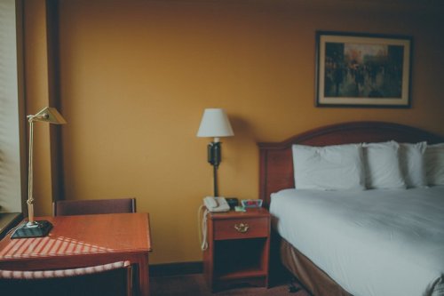 A cozy hotel room featuring a bed, desk, lamp, and a window with soft lighting.