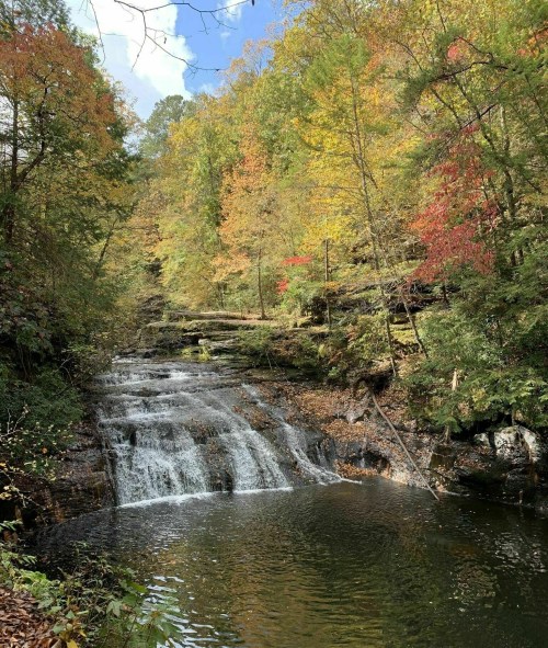 A serene waterfall cascades into a calm pool, surrounded by vibrant autumn foliage and a clear blue sky.