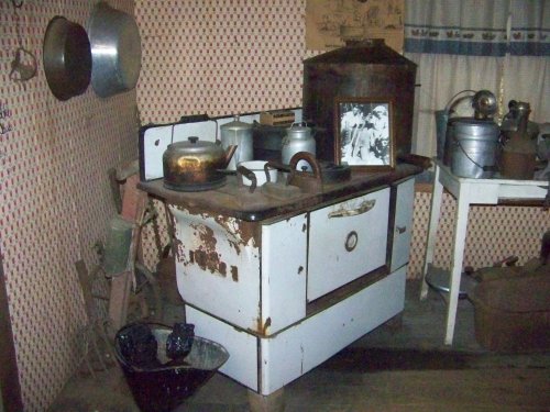 A vintage kitchen featuring an old stove, kettle, and various utensils against a patterned wallpaper backdrop.