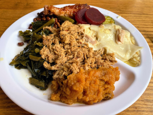 A plate of Southern-style food featuring pulled pork, collard greens, sweet potatoes, rice, and pickled beets.