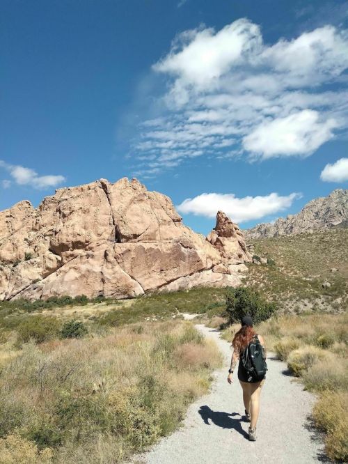 A person walks along a gravel path surrounded by rocky formations and green vegetation under a blue sky with clouds.